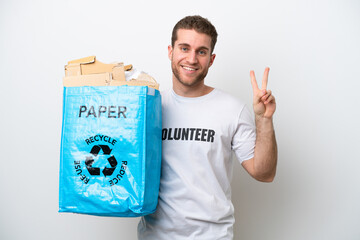 Young caucasian man holding a recycling bag full of paper to recycle isolated on white background smiling and showing victory sign