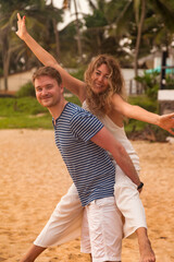 Happy lovely couple having fun on tropical sandy beach at palm trees background. Man giving piggyback ride to his woman and laughing. Joyful couple enjoying summer at ocean coastline. Copy text space