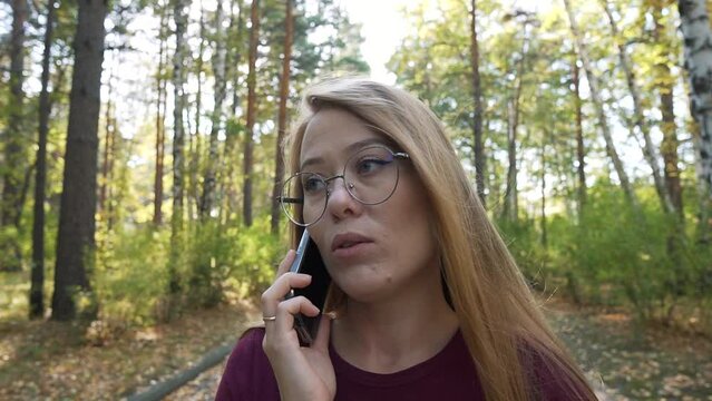 Calm woman talking smartphone in a park, telling news or gossip. Smiling girl chatting mobile phone outside. Lady walking down the path in a forest. Zoom in.