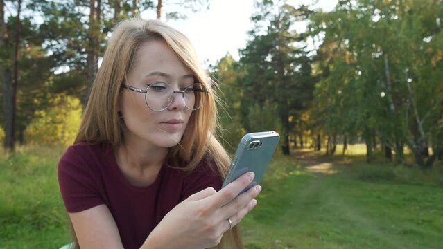 Calm woman using smartphone in a park, reading online news or swiping social media. Girl chatting in mobile phone messengers outside, sitting in a park. Zoom out shot. 