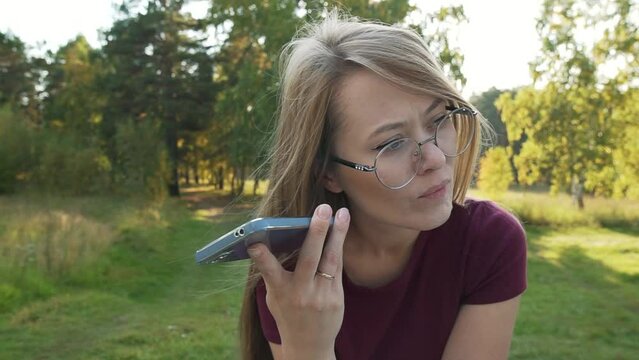 Focused woman using smartphone in a park, carefully listening audio message media from social messenger. Girl holding mobile phone sitting in a park outside. Pan shot. 
