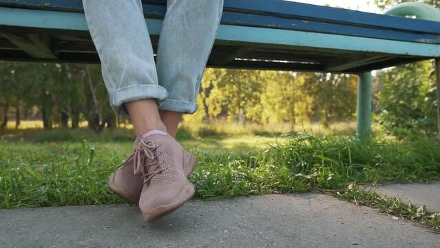 Girl dangling her feet in jeans and pink shoes. Woman swinging legs in boredom, sitting at park on a bench.
