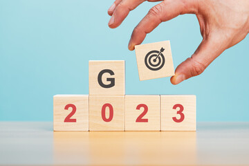 New year 2023. Hand putting wooden block with target icon on stack of wooden blocks with numbers 2023 on wooden table blue background.