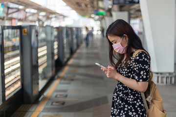Asian female  with surgical face mask  traveler looking cellphone  and typing message to her friend. she waiting for an arrival skytrain on a platform with a blurred train.