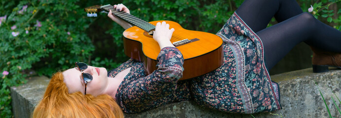 Young woman wearing sunglasses and dress lying on the stone wall in the nature and playing guitar in summer