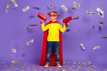 Full length photo of positive excited small son wear red superhero mantle shooting money gun isolated violet color background
