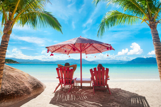 Couple on tropical beach relax in summer on deck chairs under pink umbrella. Vacation Beach Summer Holiday Concept.