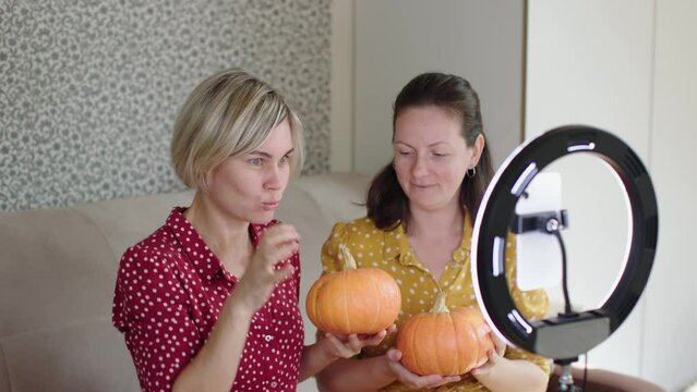 two woman bloggers filming video for social media about halloween thanksgiving holidays holding orange raw pumpkins in hands talking on camera subscribers followers. live streaming happy autumn 