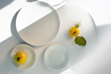 white plates of different shapes on a light background with beautiful shadows and highlights and fresh ripe quince with an empty plate inside