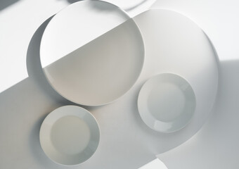 white plates of different shapes on a light background, in bright light with beautiful shadows