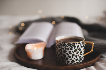 Cup of tasty coffee on wooden tray with open paper book and burning scented candle in bed close up. Cozy home hygge atmosphere. Winter season.