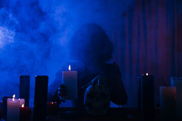 medium holding candle and touching skull during spiritual session in darkness with blue smoke.