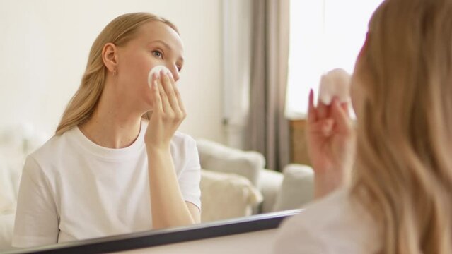 Young woman cleaning face skin with cotton pad disk and tonic. Skin care remove make up purify lotion in front mirror at home.