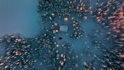 Aerial view over a cottage in middle of sunlit, winter forest of Lapland - top down, drone shot