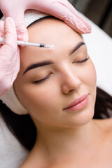 Close up of hands of young cosmetologist injecting botox in female face. She is standing and smiling. The woman is closed her eyes with relaxation