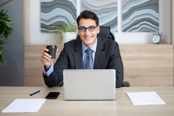 Young handsome smiling businessman working on laptop and drinking coffee in the office