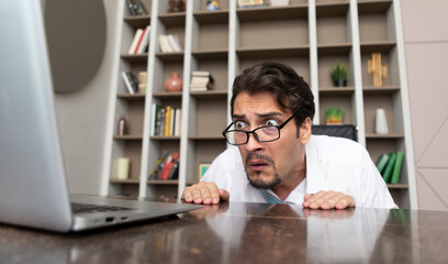 Fototapeta premium Shocked businessman hiding behind desktop and looking at laptop in the office. Business crisis and bankruptcy concept