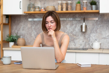 Fototapeta na wymiar Young attractive woman sitting at the table and working on laptop in the kitchen at home 