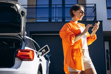Woman charging electric car and using phonr by her house