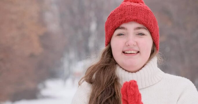 Happy cheerful teenage girl in red knitted hat and mittens looks into the camera and smiles on winter park background.
