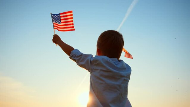 Rear view of caucasian baby boy standing and holding up his arms with two flag United States of America against blue sky. Freedom and love to country. Celebration, pride and support. Outdoor nature.