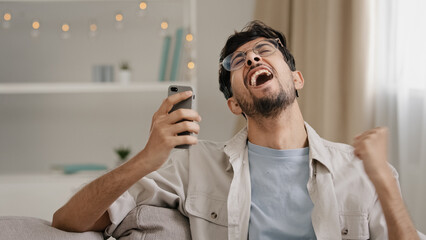 Young 30s arab bearded man millennial guy in glasses sits at home on couch looking at mobile phone making yes gesture getting offer winning good news win victory announcement feeling happy success