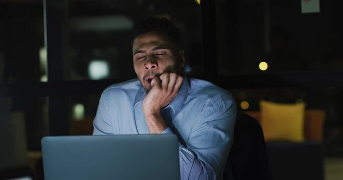 Video of tired biracial businessman sitting at desk using laptop, working at night in office