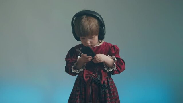Cute little girl in big headphones and a beautiful vintage dress with lace is listening to music on her phone. There is a color music in the background. The concept of music mania and audiophilia
