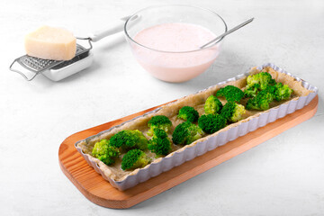 Shortbread pastry dough in rectangular pan and broccoli florets put into it. Step by step cooking tart with vegetable. Cheese and egg and cream filling in the background