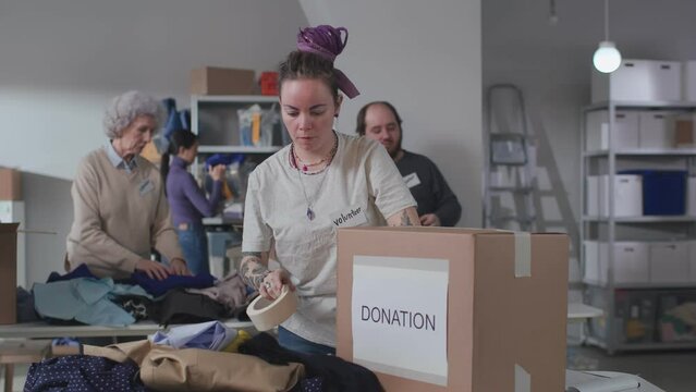 Young woman volunteer with tattoos use tape packing donation boxes in military warehouse