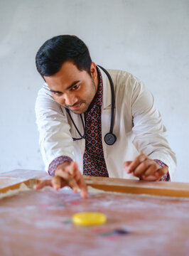 South asian young doctor passing his leisure time by playing carom board, Bangladeshi medical student wearing apron and stethoscope playing indoor game