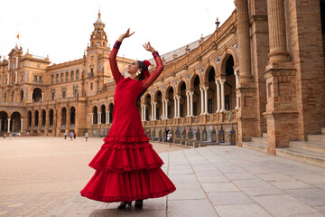 Naklejka premium Beautiful teenage woman dancing flamenco in a square in Seville, Spain. She wears a red dress with ruffles and dances flamenco with a lot of art. Flamenco cultural heritage of humanity.