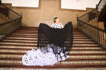 Beautiful teenage woman dancing flamenco with white dress and black polka dots doing flamenco postures on a staircase. She wears a black shawl with fringes. Flamenco cultural heritage of humanity.