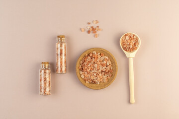 A wooden spoon and glass bootles with pink salt for body treatment or spa procedures lying on a beige background