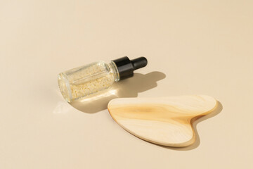 A dropper bottle with transparent antiaging serum with golden parts and a wooden guasha massager lying on a beige background