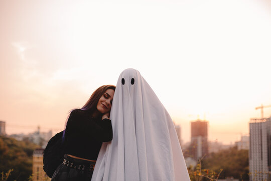 Happy young woman leaning on ghost standing against evening city during Halloween at sunset