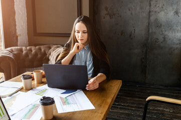 Office work. Young businesswoman looks at the laptop screen while sitting at the table. On the table are documents and glasses of coffee after a morning meeting