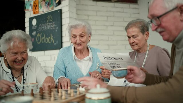 Happy senior friends sitting indoors in cafe drinking coffee or tea, playing chess laughing and having nice time together.