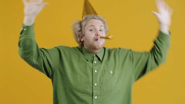 Portrait of happy man wearing birthday hat blowing horn having fun standing alone on yellow color background and looking at camera. Holidays and people concept.
