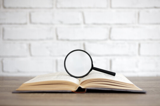 reading, education and research concept - open book with magnifying glass on the table, copy space over white brick wall background