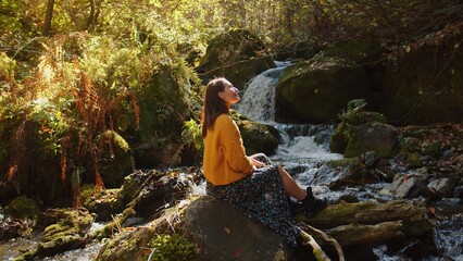 Fototapeta na wymiar Young woman is sitting on the rock nearby the cascading water stream among the autumn forest. girl enjoys the scenic landscape of colourful nature in sunny day. caucasian mountains in fall season.