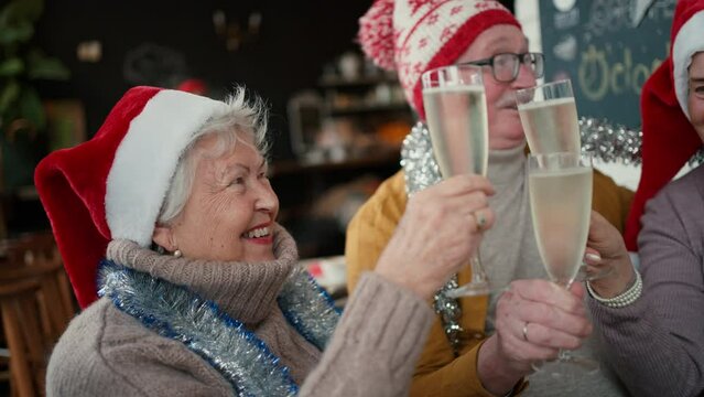 Happy senior friends sitting indoors in cafe clinking champagne glasses and celebrating Christmas.