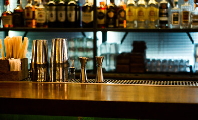 Shaker and jiger on bar counter with blurred bottles in background.