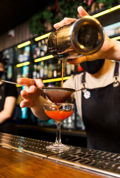 The bartender in a black protective mask pours a cocktail from a shaker into a chilled glass through a strainer. Vertical photo