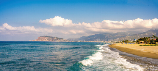 Panoramic beautiful seascape of the Mediterranean coast. The waves of the turquoise sea roll onto the sandy golden beach against the backdrop of the peninsula and mountains.