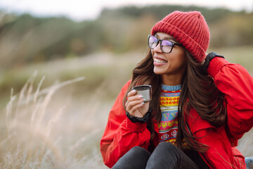 Beautiful woman drinking tea from thermos on a cold autumn day. Rest, relaxation, travel, lifestyle concept.