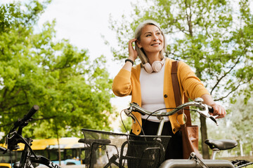 Grey asian woman with headphones standing by her bicycle at park