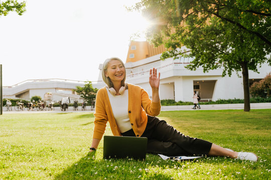 Grey Asian Woman Waving Hand And Working With Laptop On Grass