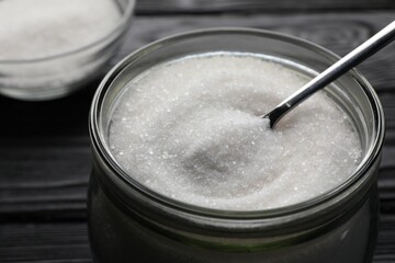 Fototapeta na wymiar Granulated sugar and spoon in glass jar on black table, closeup