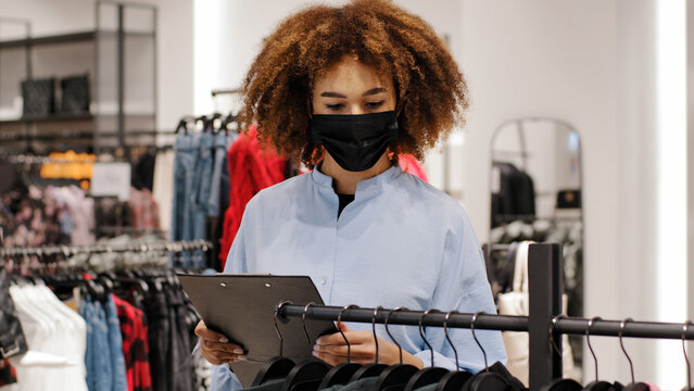 African female shop manager worker girl seller clothing store administrator make inventory salesperson woman in medical face mask owner boutique writing information about product discount size price